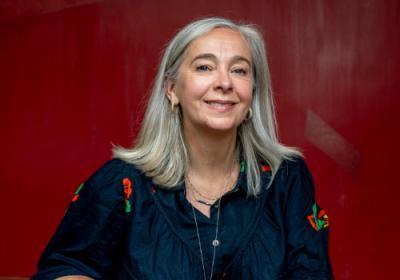 Vicky Featherstone is white woman with silver hair. She wears small hoop earrings, thin necklaces and a dark blue top.  In the background is a wall painted maroon. She has her chin up and is looking/smiling at the camera.