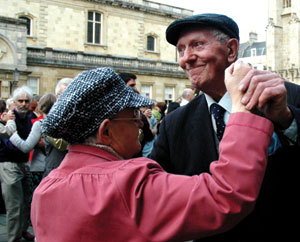 An elderly couple dance amongst a crowd of waltzing couples