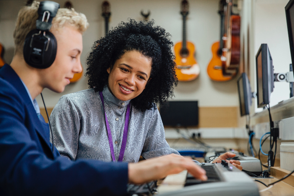 A music teacher helps a student during a music lesson. The student is playing a keyboard and the teacher is watching on and smiling