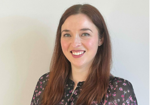 Ava Plowright has long, light brown hair. She is smiling at the camera. She has green eyes, and is wearing a black mesh shirt with pink flowers on. She stands in front of a plain, white wall.