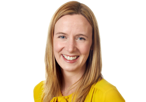 Ruth Cooke is a white woman. She has blonde hair, is wearing a yellow shirt and smiling at the camera. There is a white background behind her. Cooke is positioned to the right of the image.