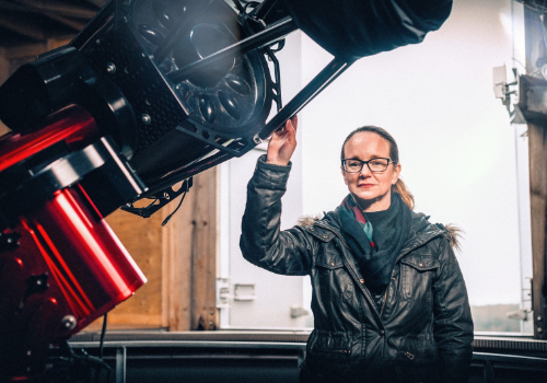 Catherine Johns is stood looking at the camera. She wears a black, leather-look puffer coat and a black scarf. She wears black glasses, and her hair is tied back. She stands in a studio and rests her hand on large stage lighting in her right hand.