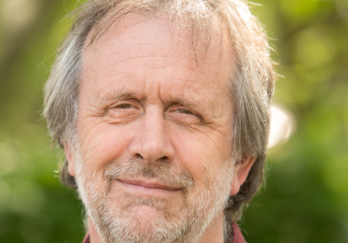 Ivan Cutting is a white man. He has grey hair and beard. He is stood outside, the trees behind him are not in focus. He is smiling and looking into the camera.