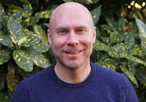 Stuart Cox is a white man. He is bald and has stubble across his chin and neck. He is wearing a dark blue jumper. In the background are green leaves.