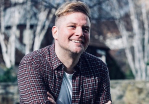 Graeme Thompson is a White man. He sits outside with his head tilted upwards and a wide smile on his face. His hair is styled to the side and he is wearing a checkered shirt with a white shirt underneath, with his arms crossed. The background is blurred.