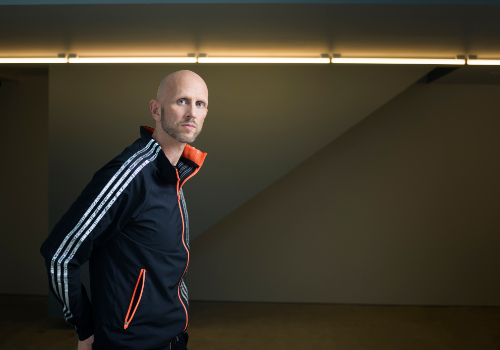 Wayne McGregor is a white man. The image is taken of his side profile, though he looks at the camera. He is bald with a short beard. He wears a tracksuit.