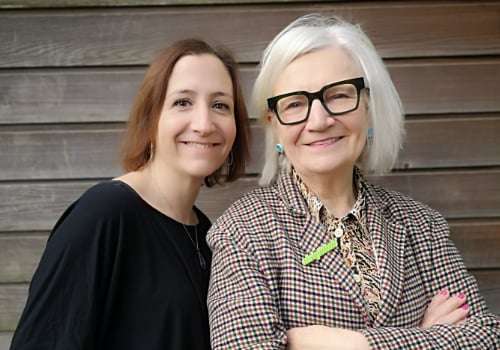 Left to Right: Jess Pearson, Janice Connolly. Jess has short brown hair and is wearing a black top. Janice has short silver hair, is wearing a checkered blazer and floral shirt. She wears black glasses, blue earrings and has her arms folded. They stand together in front of a wooden wall.