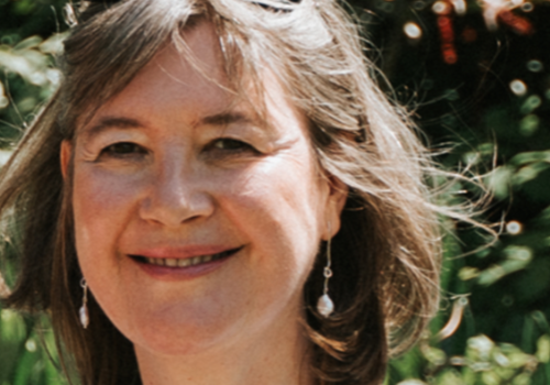 Maggie Russell is a white women with short hair. She is standing outside in the sun, wearing sunglasses on her head, pearly earrings/necklace, and a light blue collared dress. She is smiling at the camera.