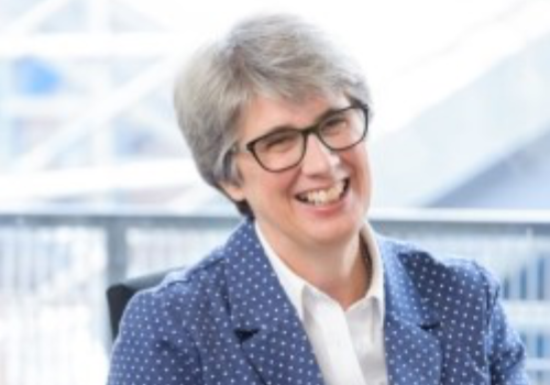 Catherine Mallyon, Trustee at Birmingham Hippodrome Theatre Trust. She is smiling, sat on a chair. Mallyon has grey, short hair and is wearing black, large glasses. She wears a blue blazer with white patterned-dots and a white shirt.