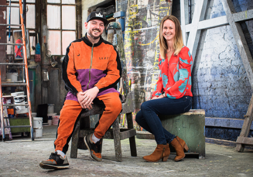 (L to R) Jesse Jones (Artistic Director), Holly Gladwell (Deputy CEO). They are photographed together in a workshop sitting on stools. Jones is a white man wearing an orange, black and purple tracksuit, black cap and trainers. Gladwell is a white woman, wearing a red and blue shirt, dark blue jeans and brown boots. They are both smiling at the camera.