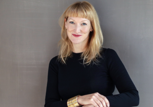 Emma Ridgway, Foundling Museum's incoming Director. She is photographed sat down against a grey background, wearing a black dress and gold bracelet. She has shoulder-length blonde hair and a short, full-fringe. She wears bright pink lipstick.