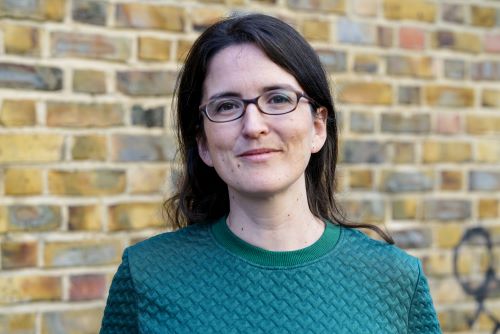 Headshot of Daisy Heath, Executive Director at Kiln Theatre. She is photographed in front of a brick wall. She wears a green top and small, black glasses. She has dark brown hair and smiles at the camera.