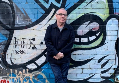 Phillip Flood stands infront of graffiti on a brick wall