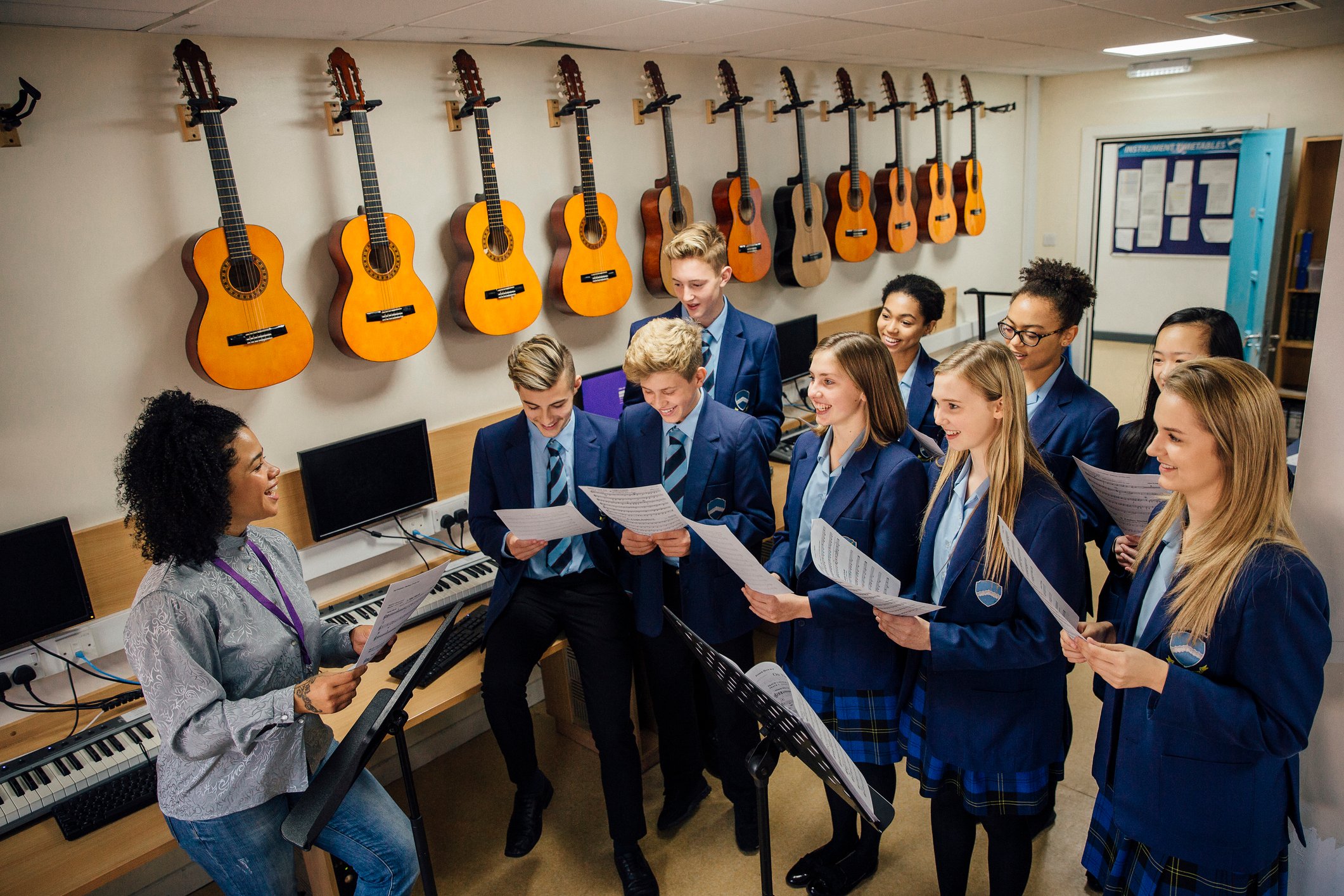 Class of teens are in their music lesson at school. There is a female teacher and the class are practicing choir with her.